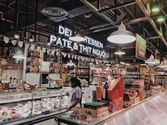 An indoor market scene featuring a deli section with a variety of meats and cheeses on display. The area is well-lit with hanging lights and has signage for 'Delicatessen' displayed prominently. Shelves are stocked with assorted goods, and a person is looking at the display counter.