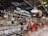 An indoor market scene featuring a deli section with a variety of meats and cheeses on display. The area is well-lit with hanging lights and has signage for 'Delicatessen' displayed prominently. Shelves are stocked with assorted goods, and a person is looking at the display counter.