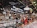An indoor market scene featuring a deli section with a variety of meats and cheeses on display. The area is well-lit with hanging lights and has signage for 'Delicatessen' displayed prominently. Shelves are stocked with assorted goods, and a person is looking at the display counter.
