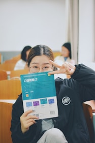 A person is holding a book titled 'College English Creative Communication' in front of their face, while making a peace sign gesture with their hand. The setting appears to be a classroom with wooden desks and several people in the background, slightly blurred.