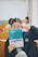 A person is holding a book titled 'College English Creative Communication' in front of their face, while making a peace sign gesture with their hand. The setting appears to be a classroom with wooden desks and several people in the background, slightly blurred.