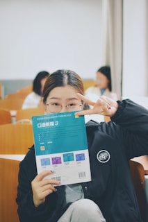 A person is holding a book titled 'College English Creative Communication' in front of their face, while making a peace sign gesture with their hand. The setting appears to be a classroom with wooden desks and several people in the background, slightly blurred.