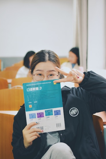 A person is holding a book titled 'College English Creative Communication' in front of their face, while making a peace sign gesture with their hand. The setting appears to be a classroom with wooden desks and several people in the background, slightly blurred.