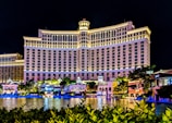A large, well-lit hotel building with an impressive facade and multiple windows overlooking a body of water. The area is adorned with greenery and small structures, all illuminated with lights, creating a vibrant scene.