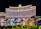 A large, well-lit hotel building with an impressive facade and multiple windows overlooking a body of water. The area is adorned with greenery and small structures, all illuminated with lights, creating a vibrant scene.