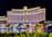 A large, well-lit hotel building with an impressive facade and multiple windows overlooking a body of water. The area is adorned with greenery and small structures, all illuminated with lights, creating a vibrant scene.