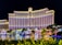 A large, well-lit hotel building with an impressive facade and multiple windows overlooking a body of water. The area is adorned with greenery and small structures, all illuminated with lights, creating a vibrant scene.
