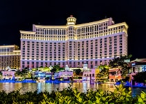 A large, well-lit hotel building with an impressive facade and multiple windows overlooking a body of water. The area is adorned with greenery and small structures, all illuminated with lights, creating a vibrant scene.