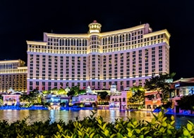 A large, well-lit hotel building with an impressive facade and multiple windows overlooking a body of water. The area is adorned with greenery and small structures, all illuminated with lights, creating a vibrant scene.