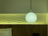A modern bedroom wall features a decorative panel with wave-like patterns. A round, white pendant light hangs in front, casting a soft glow. The bed is partially visible with a white pillow, and the color palette includes calming greens and warm wood tones.