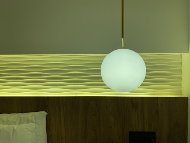 A modern bedroom wall features a decorative panel with wave-like patterns. A round, white pendant light hangs in front, casting a soft glow. The bed is partially visible with a white pillow, and the color palette includes calming greens and warm wood tones.