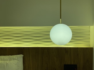 A modern bedroom wall features a decorative panel with wave-like patterns. A round, white pendant light hangs in front, casting a soft glow. The bed is partially visible with a white pillow, and the color palette includes calming greens and warm wood tones.