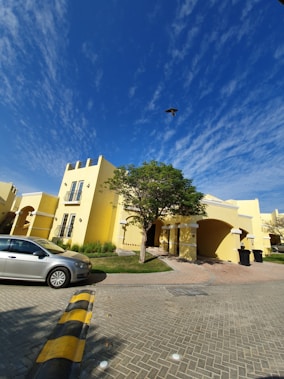 A bright, sunny day with a clear blue sky scattered with thin clouds. A modern, yellow two-story house with arched doorways and balconies stands prominently. In front, a silver car is parked on a paved driveway, and a lush green tree offers shade to part of the walkway. A bird is seen flying high above the house.