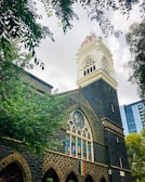 A Gothic-style building with a prominent bell tower featuring ornate architectural details, surrounded by lush green trees. A stained glass window and a rainbow flag are visible on the building's facade. The sky is overcast, with light filtering through the clouds.