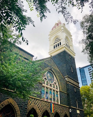 A Gothic-style building with a prominent bell tower featuring ornate architectural details, surrounded by lush green trees. A stained glass window and a rainbow flag are visible on the building's facade. The sky is overcast, with light filtering through the clouds.