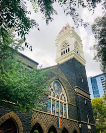 A Gothic-style building with a prominent bell tower featuring ornate architectural details, surrounded by lush green trees. A stained glass window and a rainbow flag are visible on the building's facade. The sky is overcast, with light filtering through the clouds.