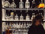 A dimly lit bar shelf is stocked with various unique bottles, some with handwritten labels in different languages. The design of the bottles varies, and they contain liquids of different colors. A person with long hair is positioned on the right side of the frame, partially in shadow.