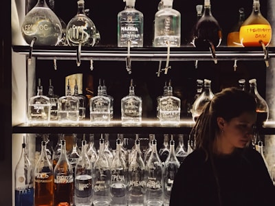 A dimly lit bar shelf is stocked with various unique bottles, some with handwritten labels in different languages. The design of the bottles varies, and they contain liquids of different colors. A person with long hair is positioned on the right side of the frame, partially in shadow.
