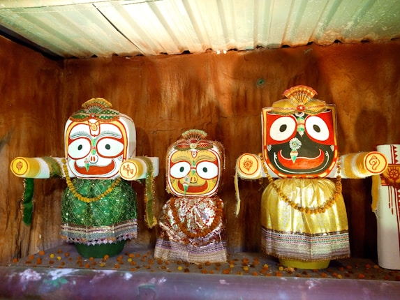 The image features three colorful, traditional Jagannath deities with large eyes, intricate patterns, and vibrant clothing. They are made of wood and embellished with beads and fabric, set against a textured brown background.