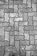An arrangement of interlocking concrete pavement blocks, featuring a repetitive zigzag pattern. The surface of the blocks shows signs of wear and has varying shades of gray, with some visible dirt and small patches of discoloration.