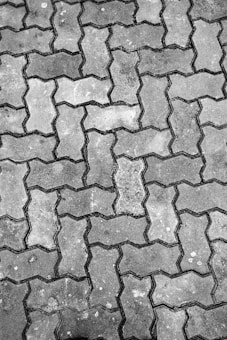 An arrangement of interlocking concrete pavement blocks, featuring a repetitive zigzag pattern. The surface of the blocks shows signs of wear and has varying shades of gray, with some visible dirt and small patches of discoloration.