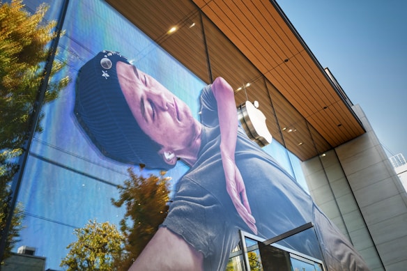 A large advertisement with an image of a person wearing a beanie and earbuds is displayed prominently on the glass facade of a modern building. The Apple logo is visible in the background above the entrance. The reflection of trees and a clear blue sky can be seen in the glass, adding a natural element to the urban setting.