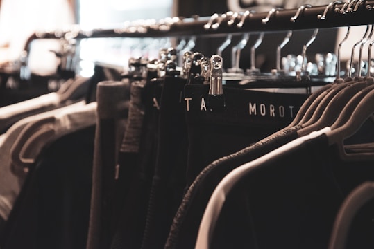 A clothing rack filled with various garments hangs neatly on metal hangers. The lighting is soft and focused, creating a warm, cozy ambiance. The clothing appears to be dark, with shades of black, gray, and possibly dark green.