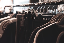 A clothing rack filled with various garments hangs neatly on metal hangers. The lighting is soft and focused, creating a warm, cozy ambiance. The clothing appears to be dark, with shades of black, gray, and possibly dark green.