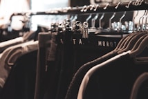 A clothing rack filled with various garments hangs neatly on metal hangers. The lighting is soft and focused, creating a warm, cozy ambiance. The clothing appears to be dark, with shades of black, gray, and possibly dark green.