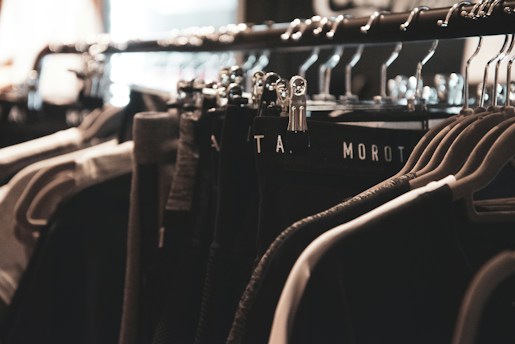 A clothing rack filled with various garments hangs neatly on metal hangers. The lighting is soft and focused, creating a warm, cozy ambiance. The clothing appears to be dark, with shades of black, gray, and possibly dark green.