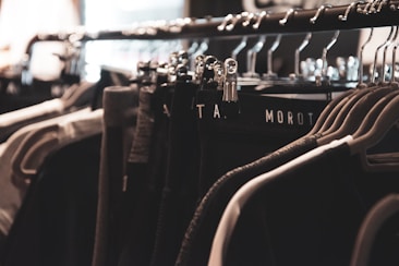 A clothing rack filled with various garments hangs neatly on metal hangers. The lighting is soft and focused, creating a warm, cozy ambiance. The clothing appears to be dark, with shades of black, gray, and possibly dark green.