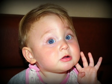 A young child with blue eyes and light brown hair is raising their hand near their face. The child appears to be wearing a light-colored bib with pink trim. The background is a dark, solid color, likely a piece of furniture or a wall.