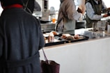 In a café or restaurant setting, there is a counter with trays containing various pastries, including a sugar-dusted cake and a pastry topped with strawberries. Several people dressed in casual clothing and aprons are engaged in activities behind the counter, possibly preparing orders.