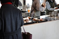 In a café or restaurant setting, there is a counter with trays containing various pastries, including a sugar-dusted cake and a pastry topped with strawberries. Several people dressed in casual clothing and aprons are engaged in activities behind the counter, possibly preparing orders.