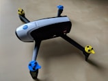 Quadcopter drone with black and white body and propellers featuring blue and yellow tops placed on a light wooden surface.