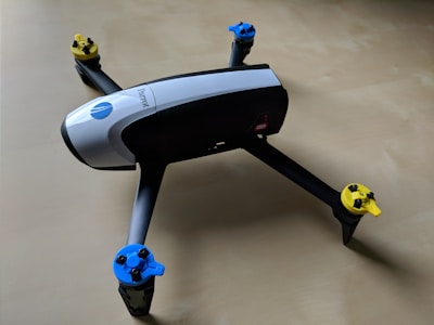 Quadcopter drone with black and white body and propellers featuring blue and yellow tops placed on a light wooden surface.