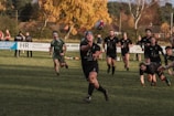 A group of rugby players is actively engaged in a match on a grassy field. One player in the foreground is passing the ball, surrounded by teammates and opponents. The players are wearing dark uniforms, some with headbands. In the background, there are spectators and autumnal trees, adding a warm color to the scene.