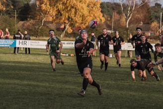 A group of rugby players is actively engaged in a match on a grassy field. One player in the foreground is passing the ball, surrounded by teammates and opponents. The players are wearing dark uniforms, some with headbands. In the background, there are spectators and autumnal trees, adding a warm color to the scene.