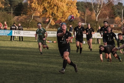 A group of rugby players is actively engaged in a match on a grassy field. One player in the foreground is passing the ball, surrounded by teammates and opponents. The players are wearing dark uniforms, some with headbands. In the background, there are spectators and autumnal trees, adding a warm color to the scene.
