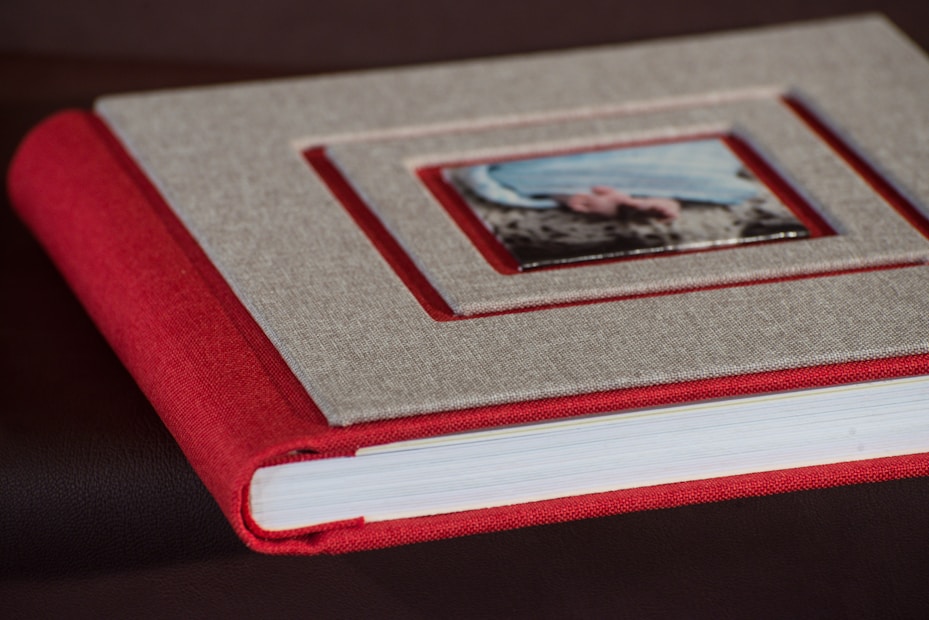 A closed photo album or book with a red spine and a textured gray cover featuring a small rectangular framed image set into the center of the front cover. The book is resting on a dark surface.