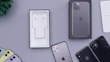 A collection of iPhones and accessories are displayed on a flat surface. The image features several iPhones in different colors, including black, yellow, and mulberry. There is an open phone box containing EarPods and a charging adapter. A box of an iPhone is positioned upright, and there's a small green plant in the corner.