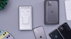 A collection of iPhones and accessories are displayed on a flat surface. The image features several iPhones in different colors, including black, yellow, and mulberry. There is an open phone box containing EarPods and a charging adapter. A box of an iPhone is positioned upright, and there's a small green plant in the corner.