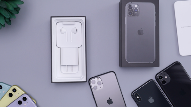 A collection of iPhones and accessories are displayed on a flat surface. The image features several iPhones in different colors, including black, yellow, and mulberry. There is an open phone box containing EarPods and a charging adapter. A box of an iPhone is positioned upright, and there's a small green plant in the corner.