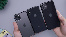 Three smartphones are being held in a person's hands, showcasing the Apple logo on their backs. The devices vary slightly in design and camera configuration. A plant and additional colorful phones are partially visible in the background.