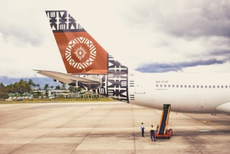 A large airplane is parked on an airport tarmac, with the tail prominently displaying intricate geometric and tribal designs in black and orange. Two ground crew members in safety vests are standing beside the aircraft, attending to operations. The sky in the background is overcast, and a line of trees and buildings is visible beyond the runway.