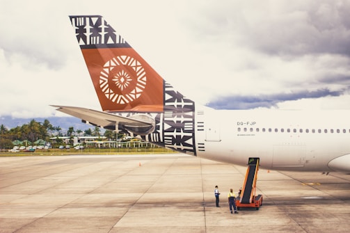 A large airplane is parked on an airport tarmac, with the tail prominently displaying intricate geometric and tribal designs in black and orange. Two ground crew members in safety vests are standing beside the aircraft, attending to operations. The sky in the background is overcast, and a line of trees and buildings is visible beyond the runway.