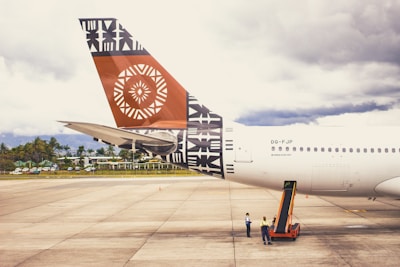 A large airplane is parked on an airport tarmac, with the tail prominently displaying intricate geometric and tribal designs in black and orange. Two ground crew members in safety vests are standing beside the aircraft, attending to operations. The sky in the background is overcast, and a line of trees and buildings is visible beyond the runway.