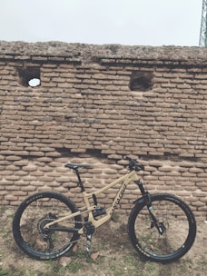 A mountain bike with a beige frame, featuring brand logos and components, is positioned against an old, rustic brick wall with a few small openings. The ground is uneven, and scattered stones and patches of grass are visible.