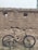 A mountain bike with a beige frame, featuring brand logos and components, is positioned against an old, rustic brick wall with a few small openings. The ground is uneven, and scattered stones and patches of grass are visible.