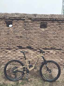 A mountain bike with a beige frame, featuring brand logos and components, is positioned against an old, rustic brick wall with a few small openings. The ground is uneven, and scattered stones and patches of grass are visible.